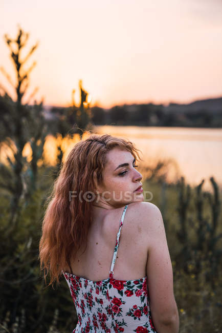 Sedutor mulher no vestido de pé na natureza no por do sol e olhando sobre o ombro — Fotografia de Stock