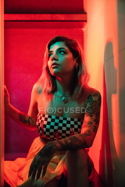 Молода жінка в одязі, дивлячись вгору, сидячи в кімнаті, освітленій червоним і зеленим світлом під час вечірки — стокове фото