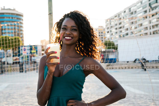 Linda mulher negra com bebida em pé na frente da rede na praia — Fotografia de Stock