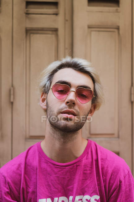 Портрет стильного чоловіка в рожевих серцеподібних сонцезахисних окулярах, що сидять навпроти бурхливих дверей — стокове фото