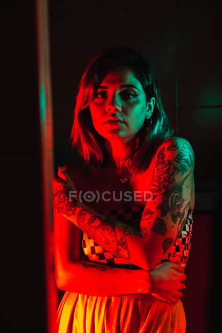 Чуттєва молода жінка з татуюваннями дивиться на камеру в темній кімнаті — стокове фото