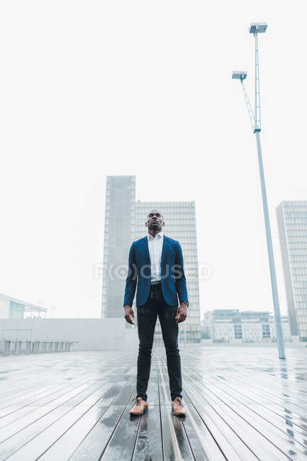 Elegante hombre de negocios de pie en el pavimento en la lluvia contra los edificios modernos de la ciudad - foto de stock