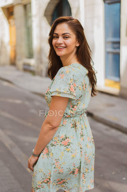 Souriant jeune femme en robe d'été marchant sur la rue étroite et regardant par-dessus l'épaule — Photo de stock