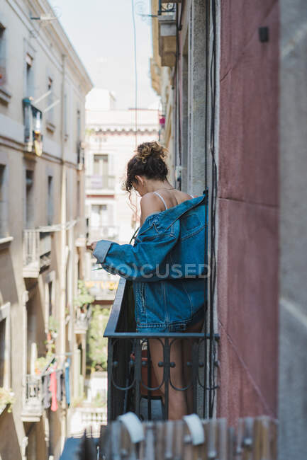 Young skinny woman in white lace underwear and denim jacket standing on small balcony with hand up looking at camera — Stock Photo