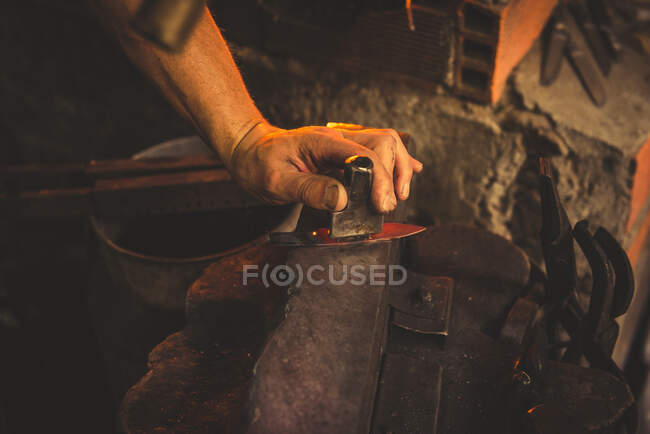 Анонимный человек использует Хаммер для ковки лезвия из куска металла в профессиональной мастерской — стоковое фото