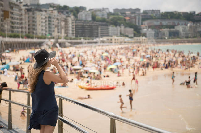 Жінка тримає капелюх і насолоджується видом на переповнений пляж і спокійне море, стоячи біля паркану — стокове фото