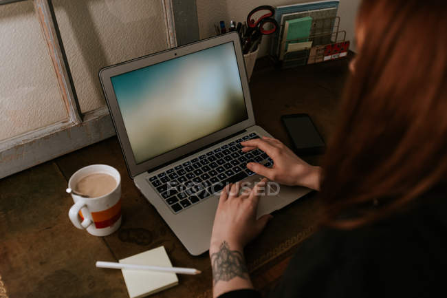Nahaufnahme einer tätowierten Frau, die am Laptop tippt und ihr Smartphone am Tisch hält — Stockfoto