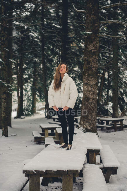 Blonde Dame in weißer Jacke posiert im Winter auf hölzernem Picknickplatz und blickt in die Kamera — Stockfoto