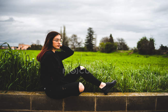 Portrait of stylish young woman sitting on brick wall in countryside in overcast — Stock Photo