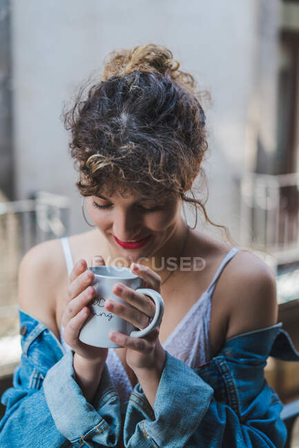 Jeune fille bouclée en sous-vêtements blancs et veste en denim debout sur le balcon avec tasse de café regardant Dreamily loin — Photo de stock
