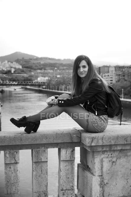 Vue latérale de la jeune femme attrayante en veste en cuir et jupe courte avec tatouage sur la jambe et sac à dos assis sur le parapet en pierre du pont à Bilbao et regardant la caméra sur le fond de la rivière — Photo de stock