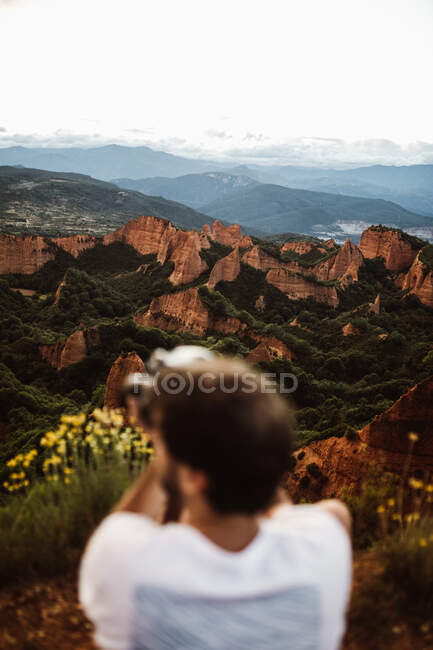 Vista posterior de fotógrafo borroso con cámara haciendo foto de pie en la cima de las colinas en el fondo del paisaje en Cantabria, España - foto de stock