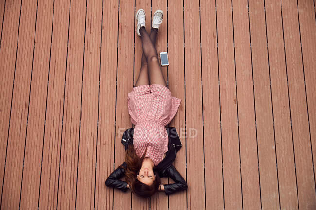 De cima vista de jovem mulher atraente em jaqueta de couro e vestido deitado na superfície listrada com telefone celular colocado perto — Fotografia de Stock