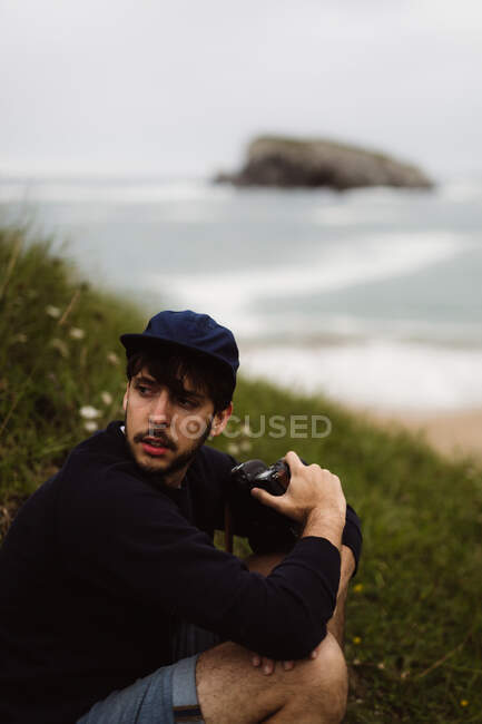 Jovem sentado na grama na costa e olhando para longe, segurando a câmera na mão e no mar no fundo na Cantábria, Espanha — Fotografia de Stock