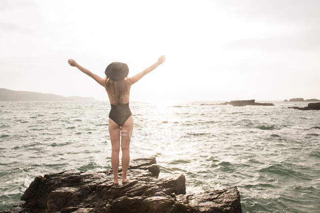 Жінка в купальнику, стоячи руками вгору на валуні в морі в сонячний день — стокове фото