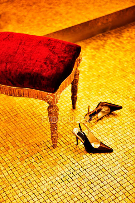 Black leather high heel shoes placed on tile floor near ancient chair with patterned legs and red cover — Stock Photo