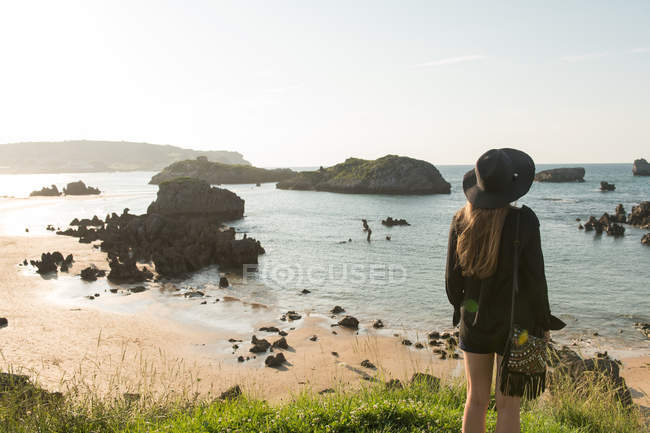 Mujer en sombrero de pie en la hierba en la costa y disfrutar de la vista del mar en el día soleado - foto de stock