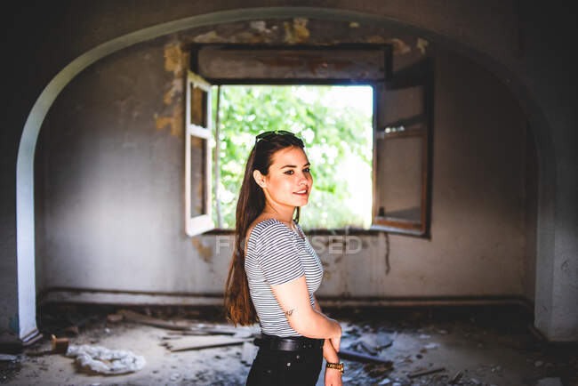 Attraente giovane donna in abito casual guardando la fotocamera mentre in piedi in corridoio di vecchio edificio abbandonato. — Foto stock