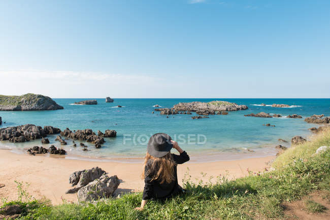 Woman in hat sitting on grass on coast and enjoying view of sea on sunny day — Stock Photo