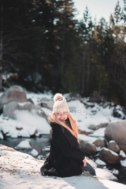 Mujer bonita sonriente sentada y mirando hacia el río en la naturaleza invernal. - foto de stock