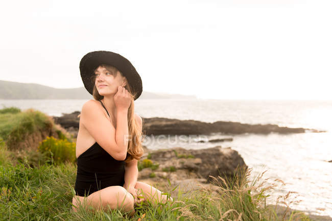 Young woman in black swimsuit and hat sitting near at sea — Stock Photo