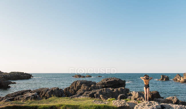 Жінка в купальнику і капелюсі стоїть на траві на узбережжі і насолоджується видом на море в сонячний день — стокове фото