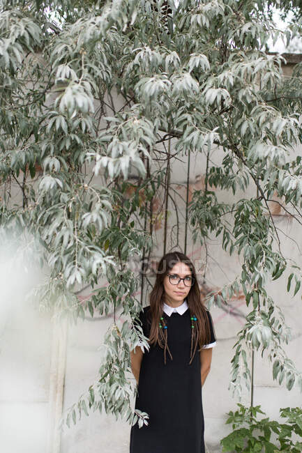 Junge charmante Frau in schwarzem Kleid und Brille steht unter grünem Baum und blickt in die Kamera — Stockfoto