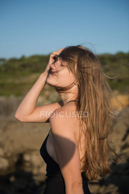 Молода жінка з закритими очима регулює волосся, стоячи на розмитому тлі природи — стокове фото