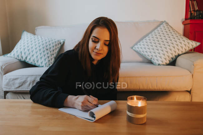Молодая женщина пишет в блокноте за столом со свечой дома — стоковое фото