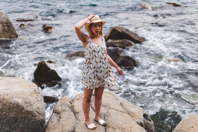 Visão traseira da mulher tatuada de chapéu em pé sobre pedras com as mãos afastadas e olhando para o oceano — Fotografia de Stock