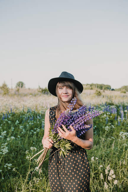 Стильная женщина в шляпе и платье, стоящая с букетом фиолетовых цветов в сельском поле, улыбается перед камерой — стоковое фото