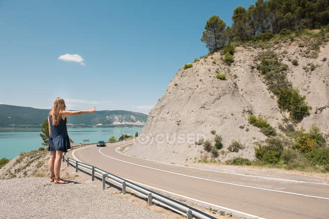 Young woman hitchhiking on roadside of narrow countryside road at sea — Stock Photo