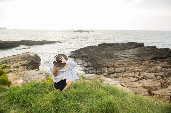 Mulher de chapéu sentado na grama na costa e desfrutando de vista para o mar — Fotografia de Stock