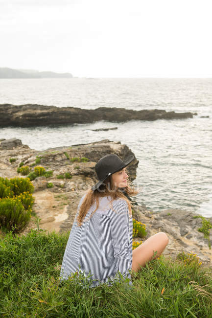 Jovem mulher de chapéu sentado na grama na costa e desfrutando de vista para o mar — Fotografia de Stock