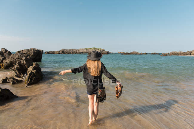 Vista trasera de la mujer en sombrero y vestido caminando en la orilla arenosa en medio de rocas hacia el mar - foto de stock