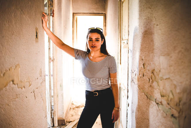 Atractiva mujer joven en traje casual mirando a la cámara mientras está de pie en el pasillo del viejo edificio abandonado. - foto de stock