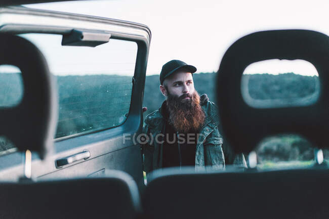 Вид сбоку на взрослого бородатого мужчину, открывающего багажник машины и отводящего взгляд в сторону на природе. — стоковое фото
