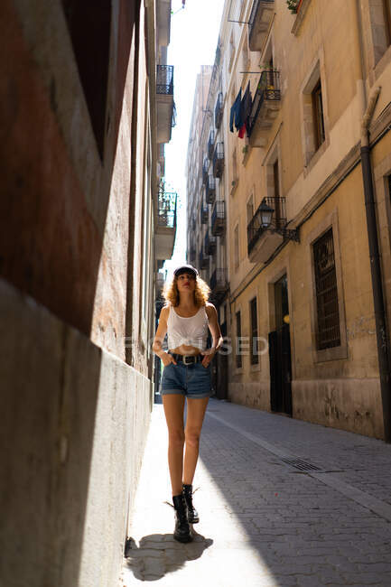 Очаровательная кудрявая женщина в шортах и жестоких сапогах, игриво стоящая на асфальтированной улице и развлекающаяся — стоковое фото