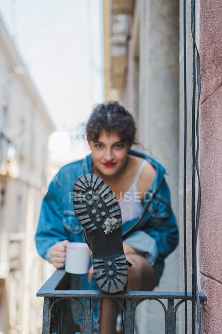 Красивая молодая девушка с кофейной чашкой, стоящая на балконе и держа ногу на заборе, показывая подошву — стоковое фото