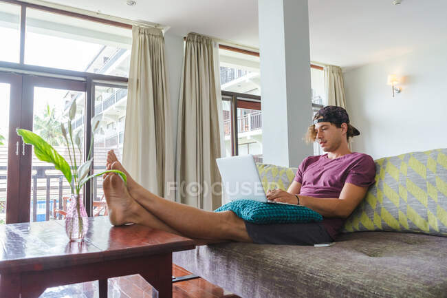 Homme adulte assis sur un canapé avec des jambes sur la table et utilisant un ordinateur portable à la maison — Photo de stock