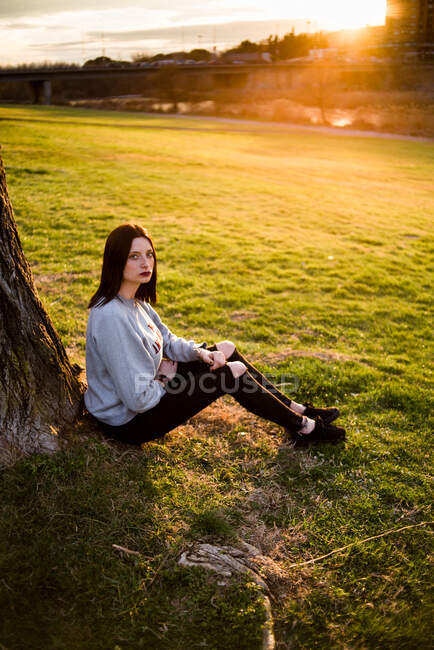 Side view of casual brunette sitting under tree on lawn against city in sunset and gloomy sky. — Stock Photo