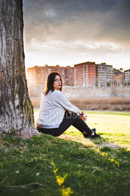 Side view of casual brunette sitting under tree on lawn against city in sunset and gloomy sky. — Stock Photo