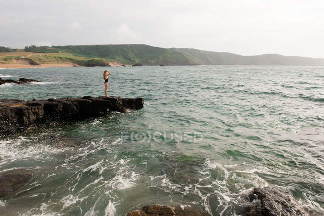 Женщина в купальнике, стоящая на скале в море и смотрящая на вид — стоковое фото