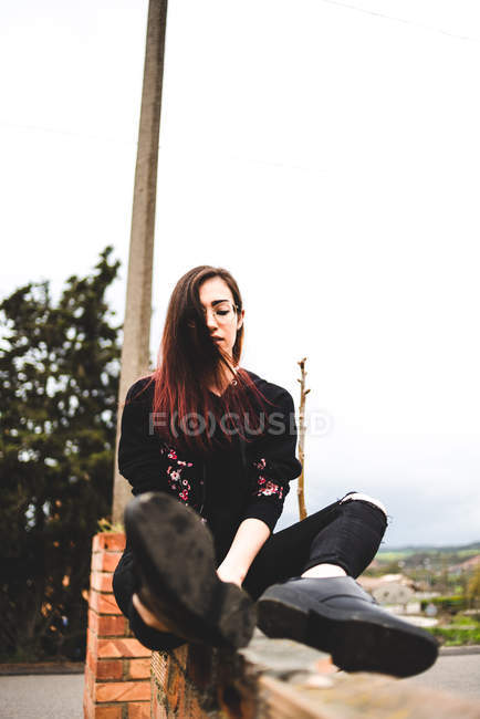 Stylish young woman with eyes closed sitting on brick wall in countryside in overcast — Stock Photo