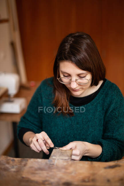 Donna concentrata in camicetta verde e occhiali decorazione intaglio con strumento al tavolo da lavoro in officina — Foto stock