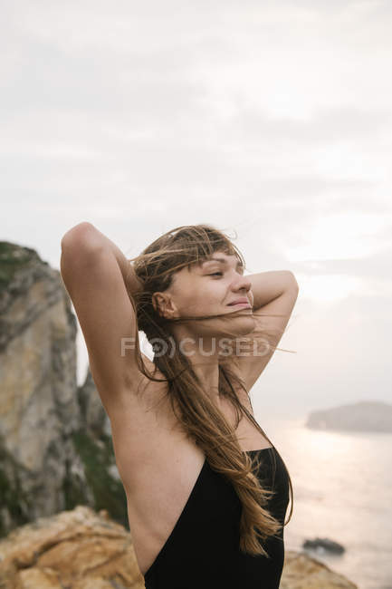 Jovem mulher com as mãos atrás da cabeça em pé na costa rochosa — Fotografia de Stock