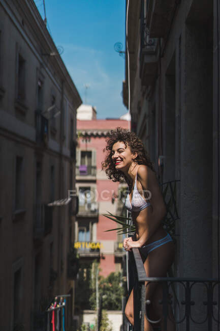 Chica encantadora en ropa interior y botas con chaqueta de pie en el balcón y sonriendo a la cámara a la luz del sol - foto de stock