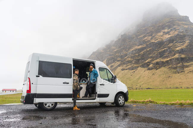 Dos jóvenes de pie cerca de una bonita furgoneta blanca mientras viajan a través de magníficos paisajes islandeses - foto de stock