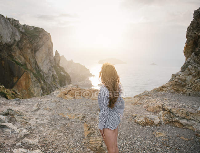 Sinnliche junge Frau im Hemd, die an der felsigen Küste steht und die Aussicht betrachtet — Stockfoto
