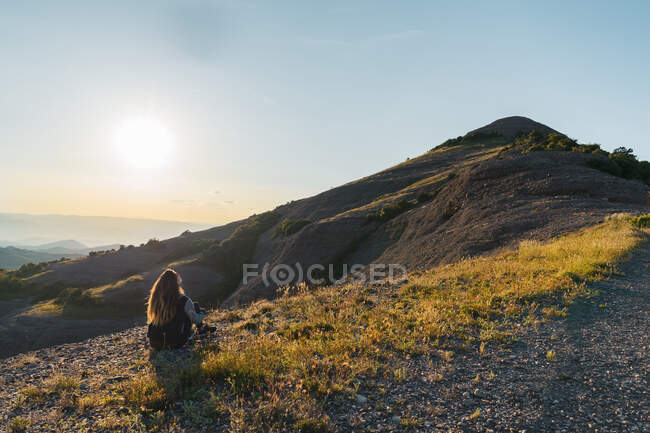 Back view of female in warm clothes sitting near top of mountain covered with rubble and short alpine grass at sunrise in Spain — Stock Photo